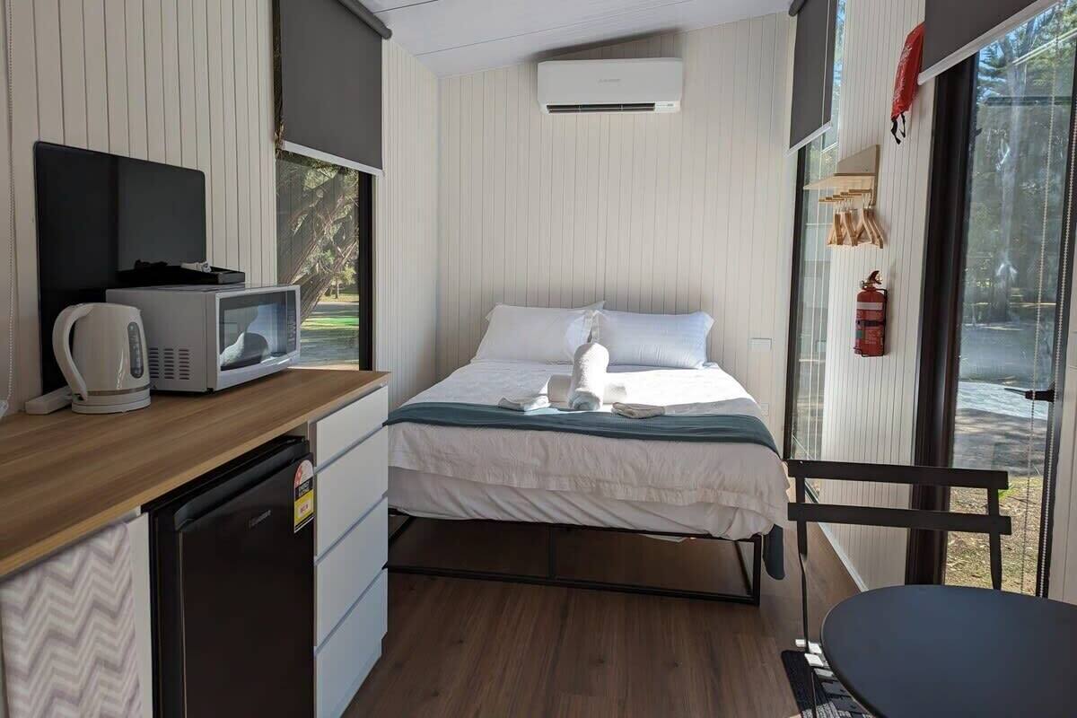 Una habitación compacta con cama, kitchenette pequeña, microondas, hervidor y extintor.