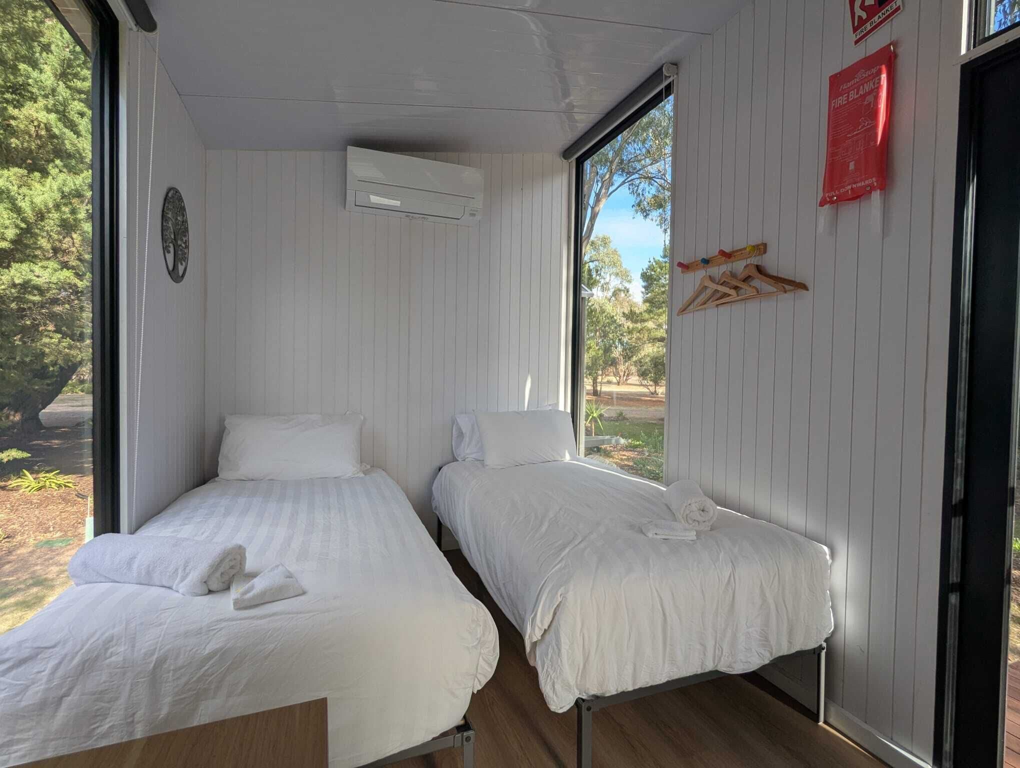 Une chambre avec deux lits simples, une fenêtre donnant sur des arbres, un appareil de climatisation et un panneau indiquant la présence d’une couverture anti-feu.