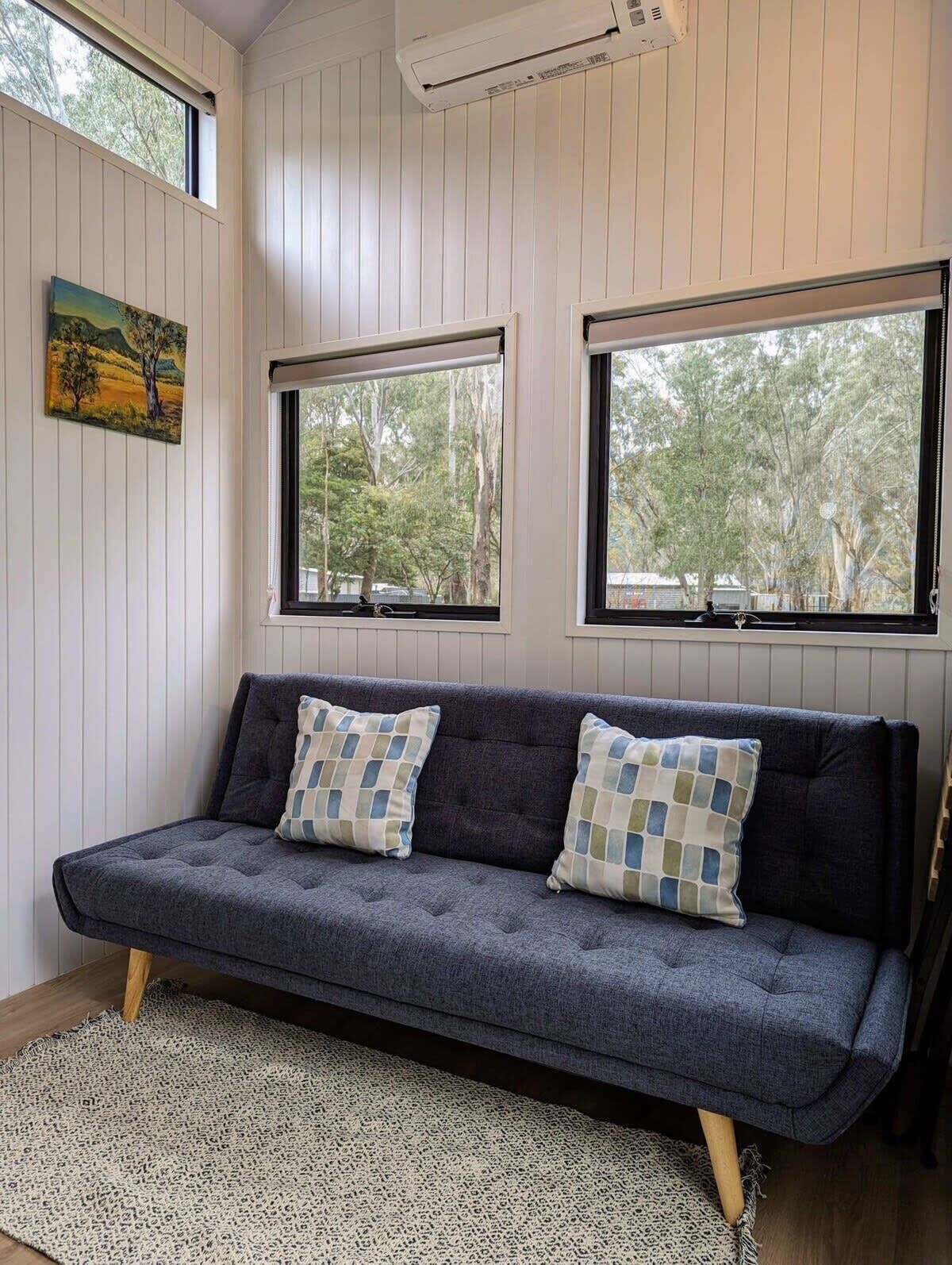 A room with a dark grey sofa, two patterned cushions, a woven rug, and a painting on the wall.