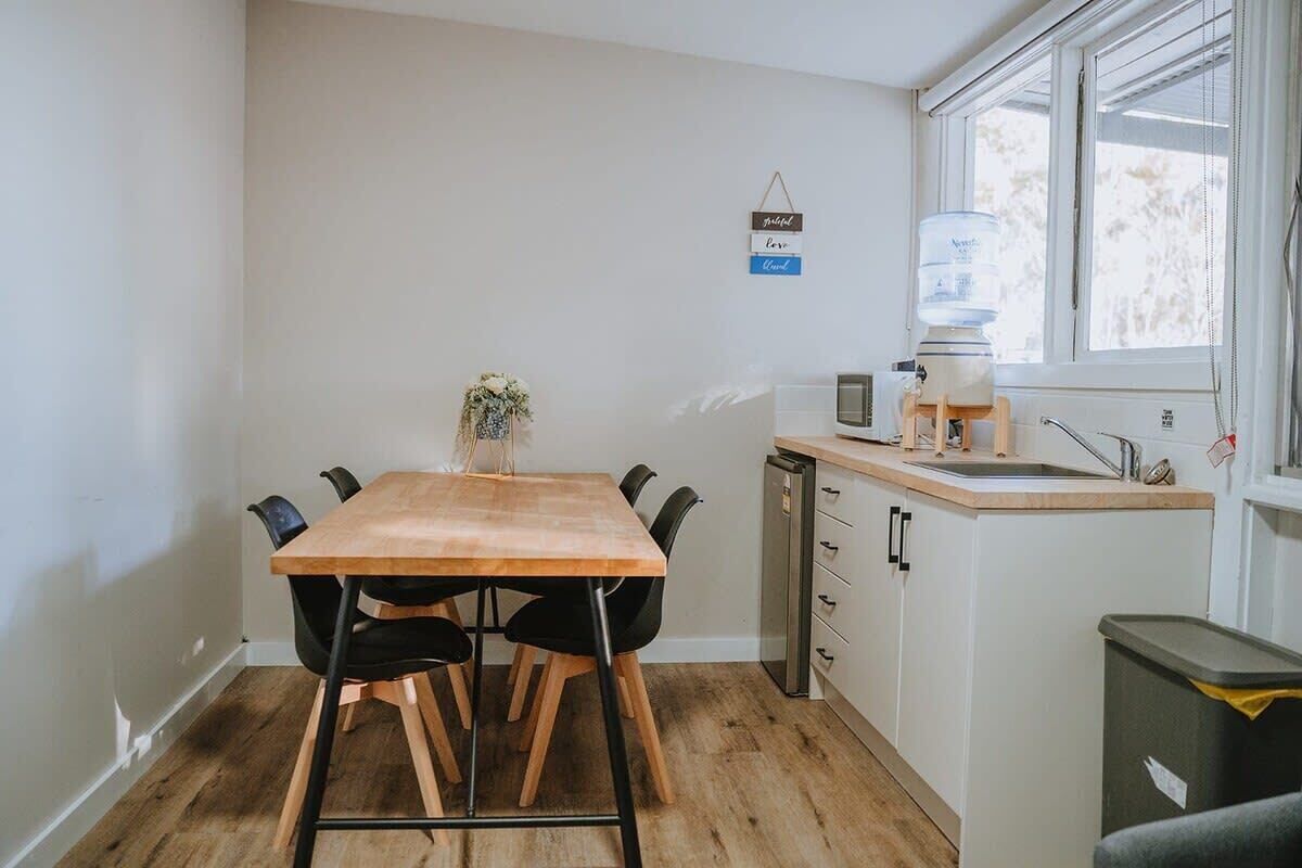 Una cocina compacta con una mesa de madera, sillas negras, un dispensador de agua y un lavamanos pequeño.