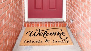 Une porte rouge dotée d’un clavier pour ouvrir la serrure et d’un paillasson sur lequel est écrit : « Bienvenue, amis et famille ».