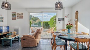 A spacious living area with a large sliding glass door leading to a garden, a cozy sofa, a dining table set for four, and a television on a wooden stand.