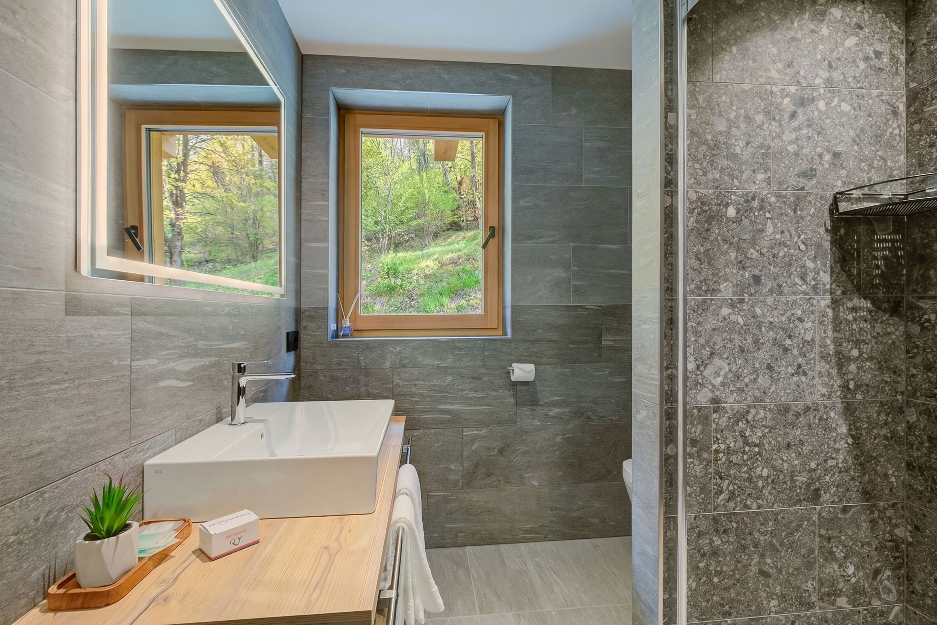 Une salle de bain moderne avec une grande baignoire, deux fenêtres donnant sur un paysage verdoyant et un mur en pierre.