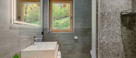 Une salle de bain moderne avec une grande baignoire, deux fenêtres donnant sur un paysage verdoyant et un mur en pierre.