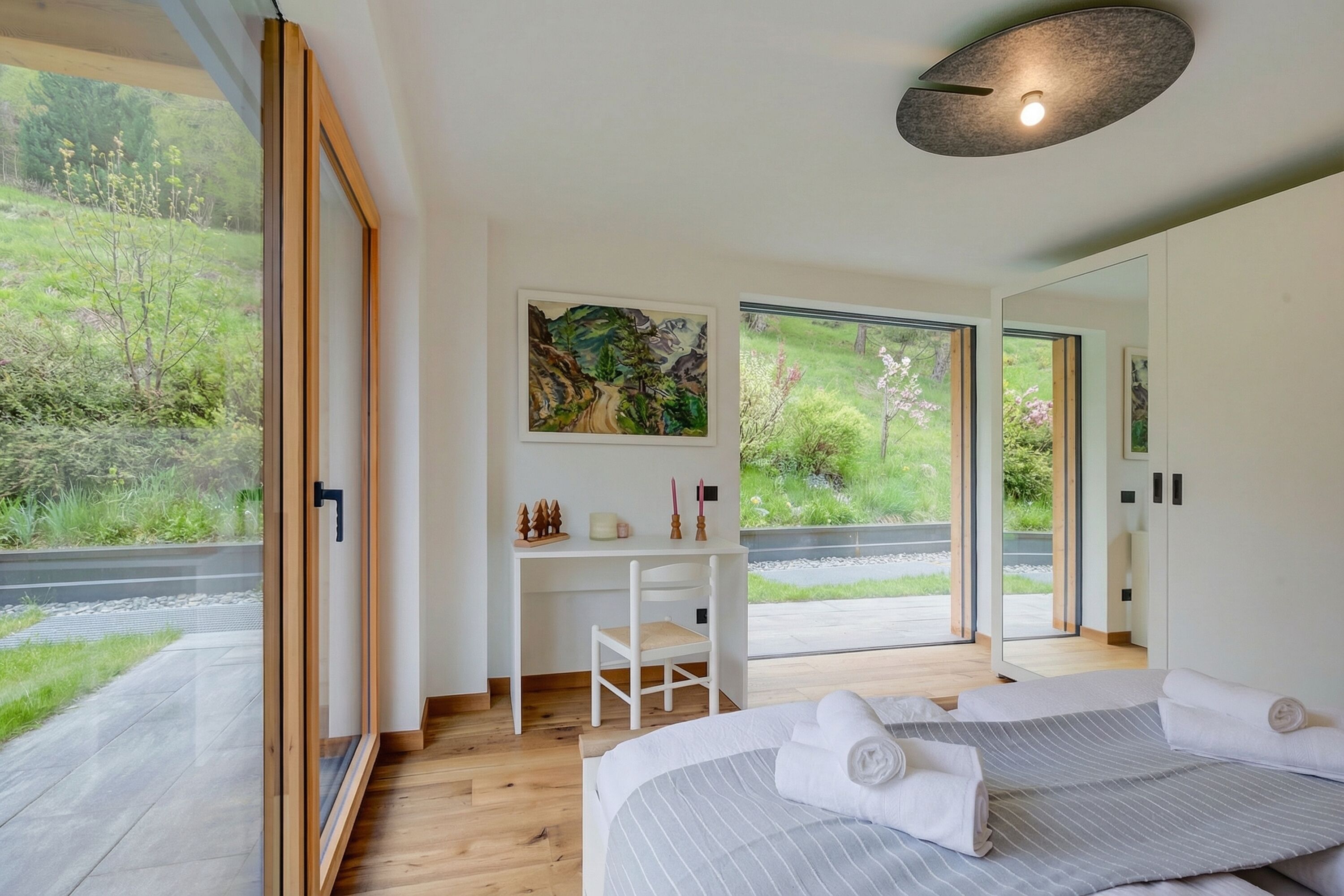 A modern bedroom with a large bed, a desk, and a chair, a painting on the wall, and a view of greenery outside.