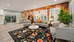Ein Wohnzimmer mit floralem Teppich, grauen Sofas, einem Couchtisch und einem großen Fenster mit Jalousien.