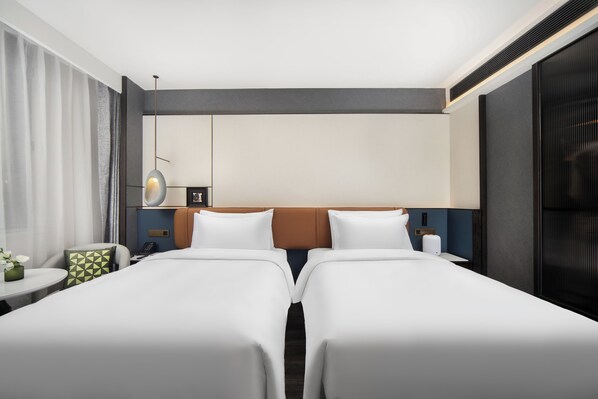 Habitación de hotel con dos camas, una mesita con un jarrón, una lámpara y vistas a la ciudad.