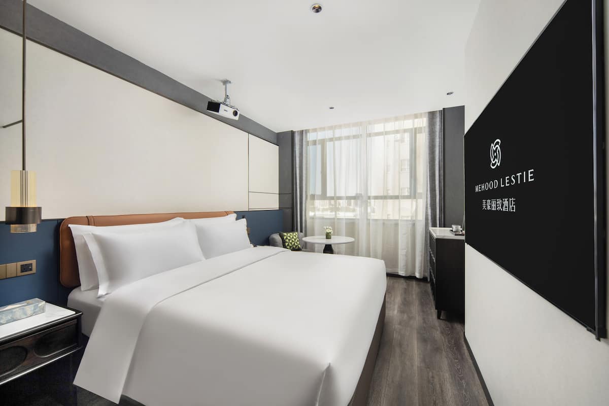 Una habitación de hotel moderna con una cama grande, una mesita de noche, una pequeña mesa redonda y una ventana con cortinas.