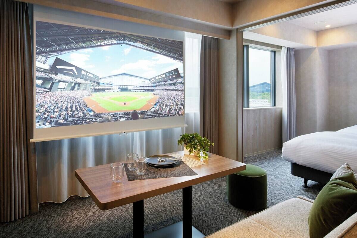 Habitación de hotel con una ventana grande, una cama, una mesita de noche con una planta y vistas a un estadio de béisbol.