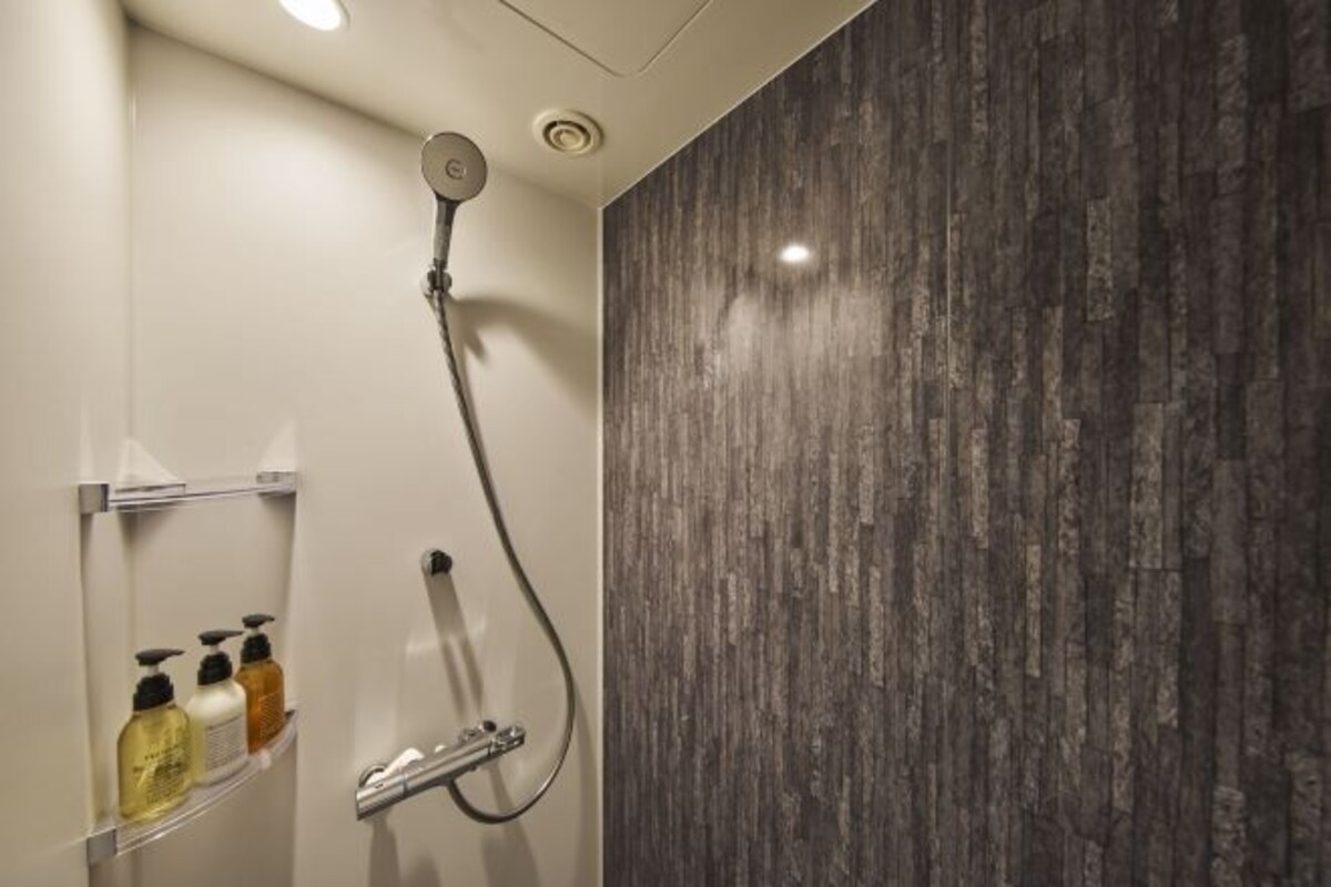 A modern shower with a rain showerhead and a handheld showerhead, a shelf with toiletries, and a textured wall.