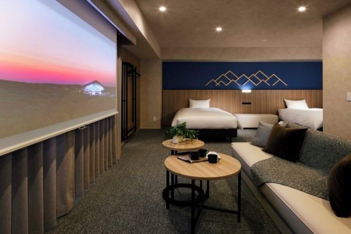 A modern hotel room with a large bed, a sofa, a small table, and a projector screen displaying a sunset view.