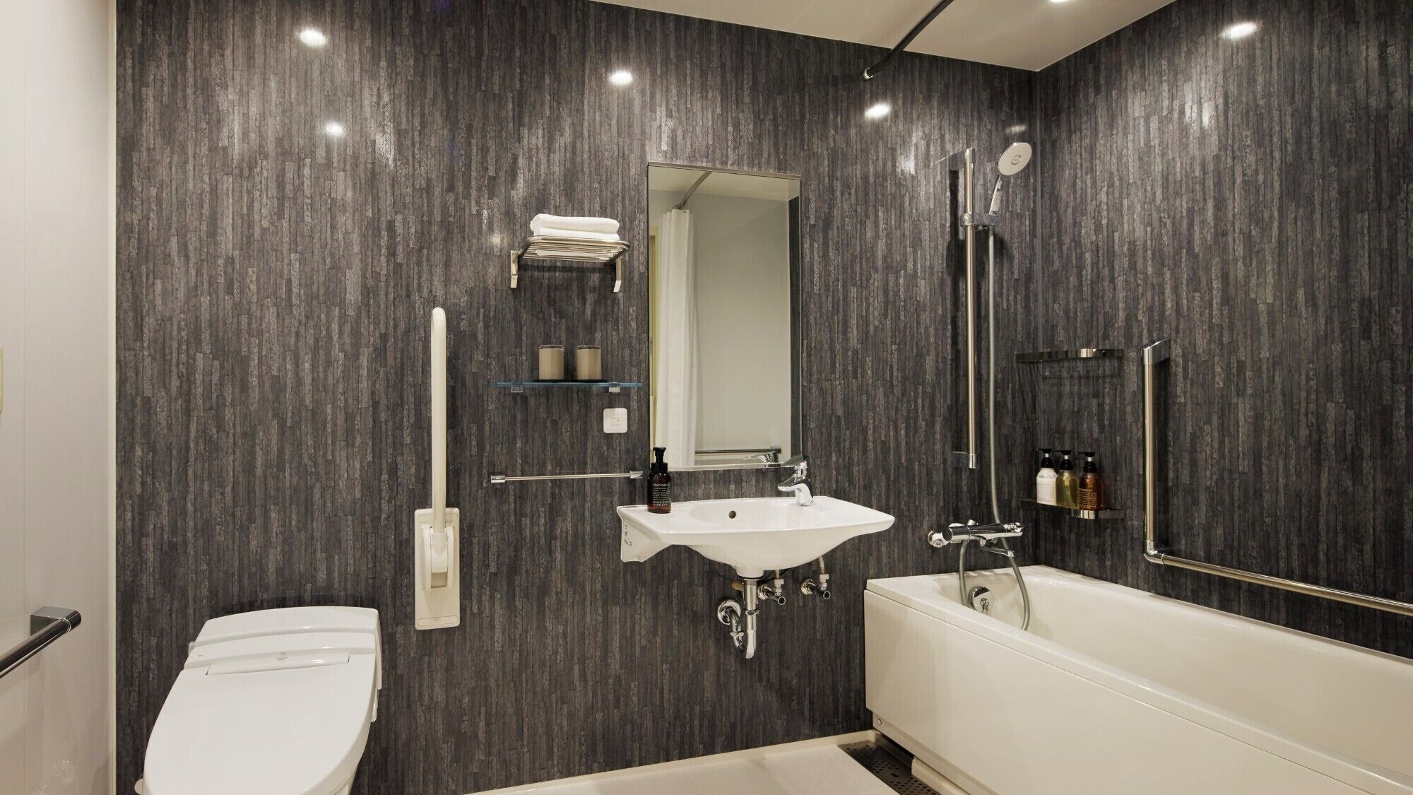 Une salle de bain moderne avec un lavabo blanc, une baignoire et des toilettes. On y trouve des étagères avec des articles de toilette et un miroir au-dessus du lavabo.