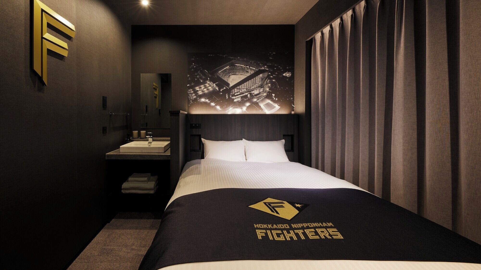 A hotel room with a bed, a nightstand, a sink, and a mirror. The bed has a black and white bedspread with the Fighters logo.