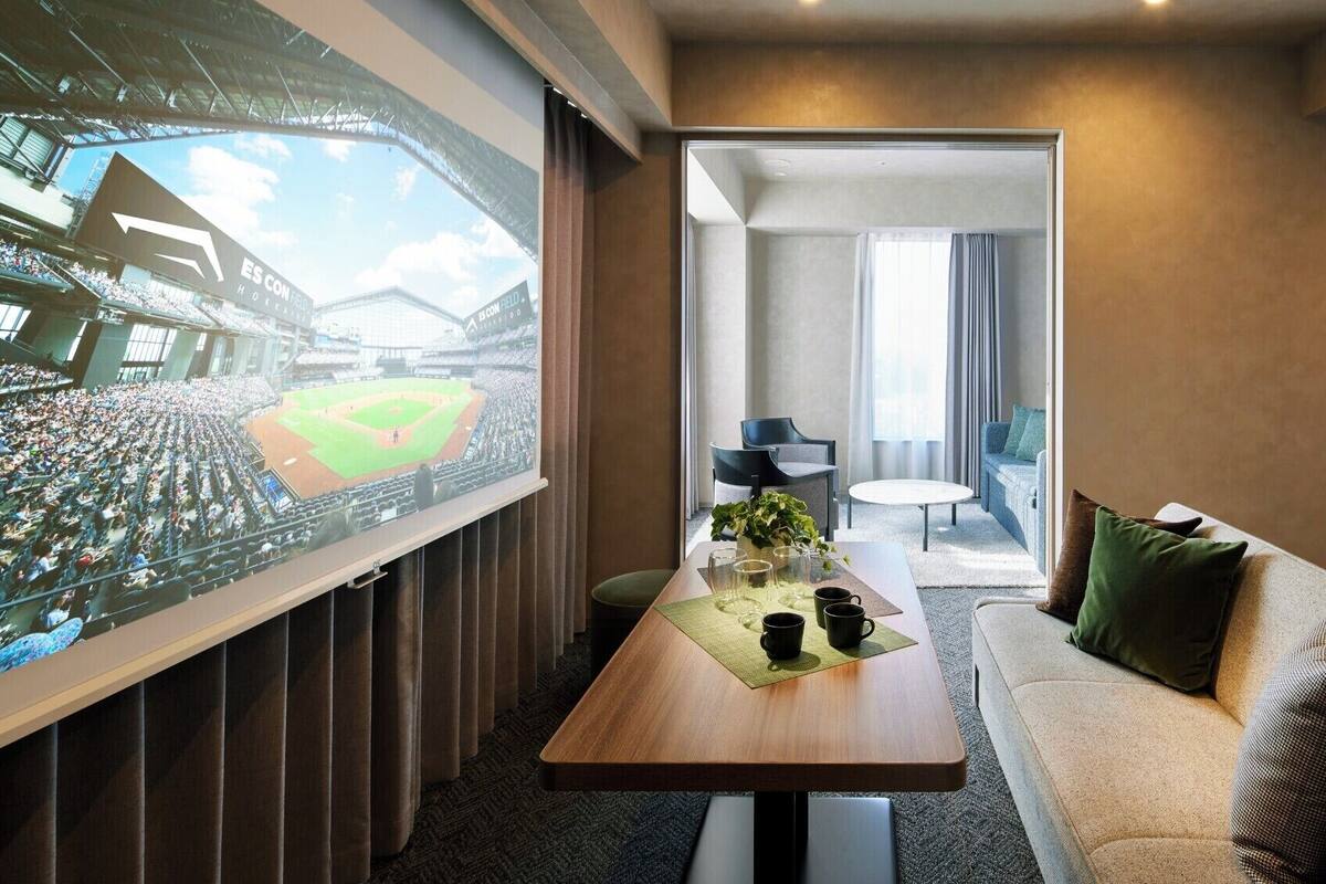 Habitación de hotel con una pantalla grande mostrando un partido de béisbol, un área de comedor con una mesa para dos, y una zona de estar acogedora con un sofá y cojines.