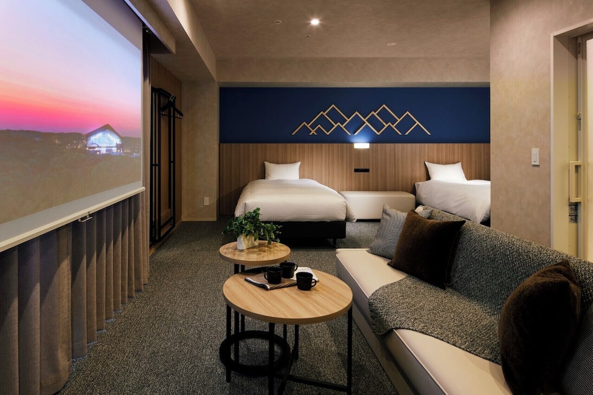 Habitación de hotel con una pantalla de proyección grande, un sofá, dos camas y una mesita con una planta.
