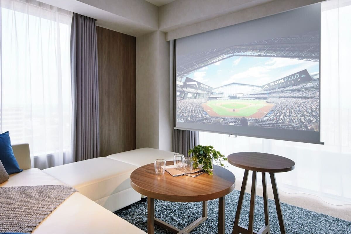 Habitación de hotel con una pantalla grande mostrando un partido de béisbol, un sofá, una mesita con vasos y una planta.