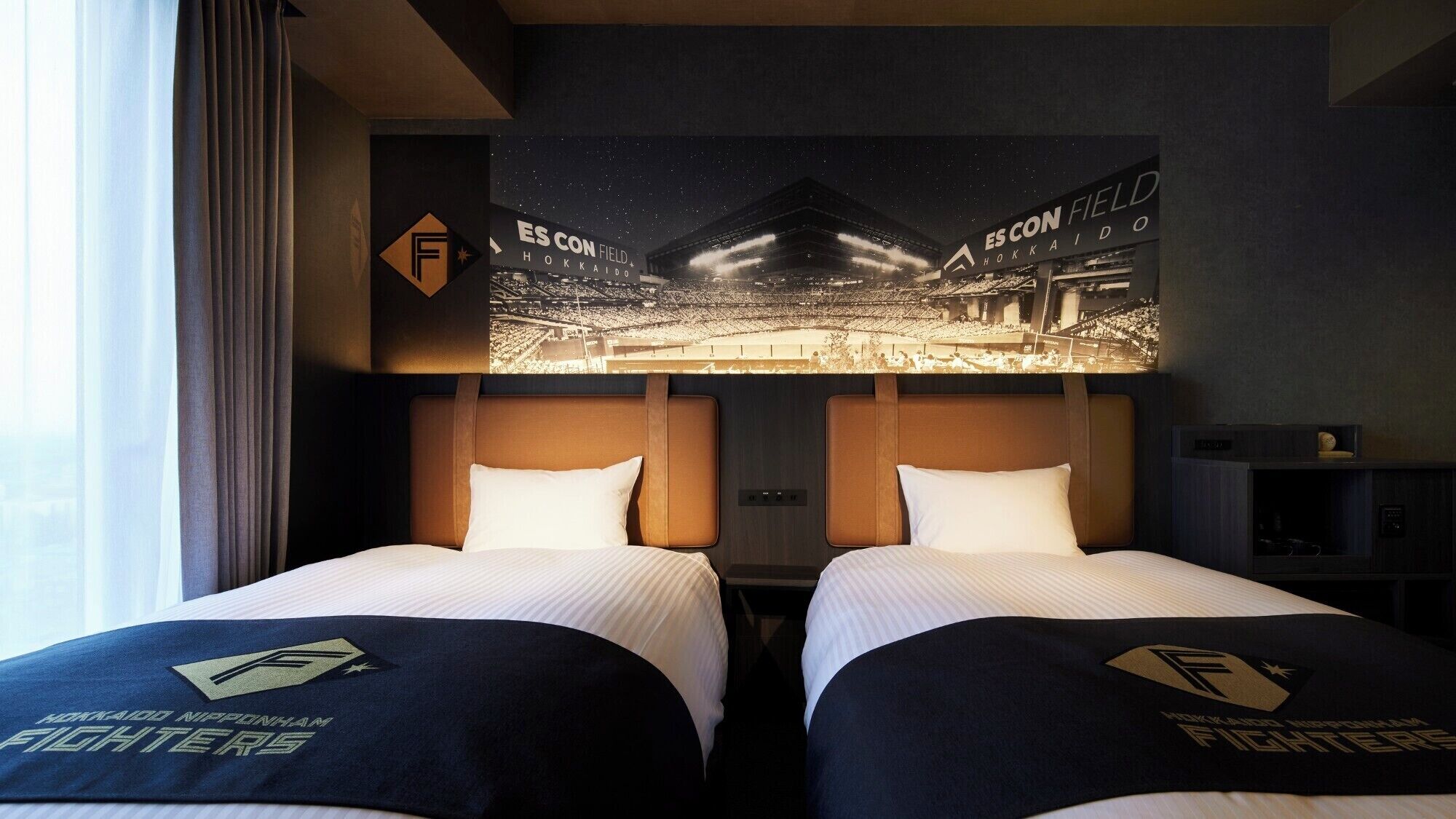 Habitación de hotel con dos camas, cada una con un almohadón que lleva un logotipo. La habitación tiene un cartel grande de un estadio en la pared, y se aprecia una vista del cielo nocturno a través de la ventana.