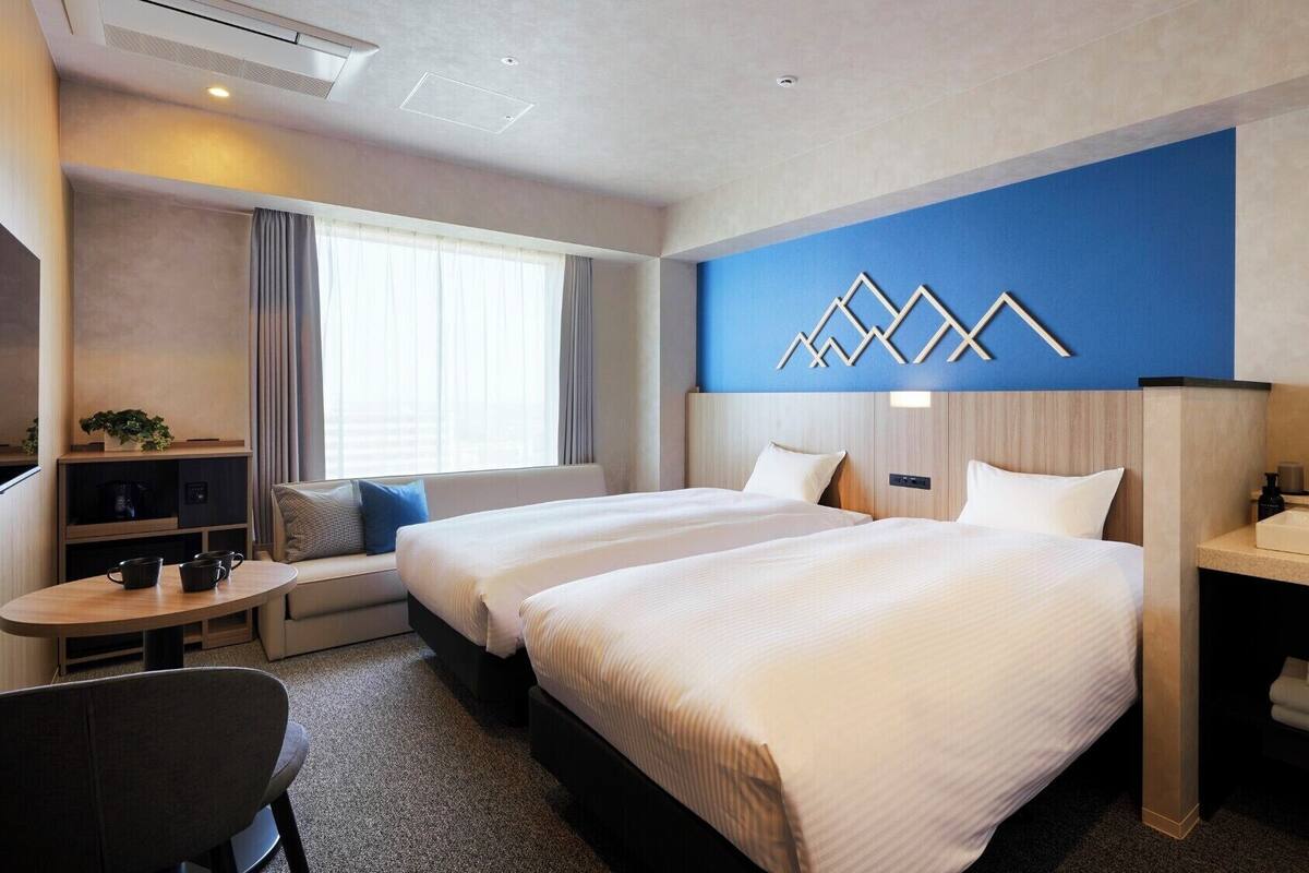 Habitación de hotel con dos camas, un escritorio, una silla, una mesita, un sofá, un ventanal grande, un espejo en la pared y un baño con lavamanos y toallero.
