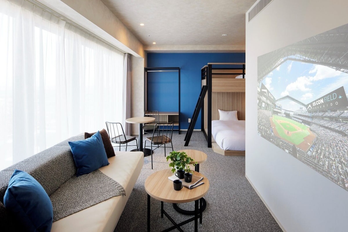Habitación de hotel con sofá, una mesita con una planta, literas y un ventanal con cortinas de encaje.