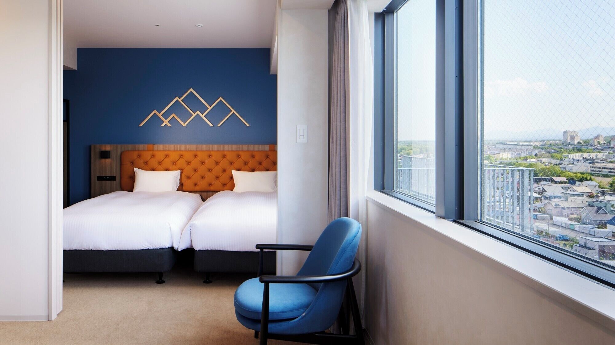 Ett hotellrum med ett stort fönster, två sängar, en blå stol och en dekorativ väggdekoration.