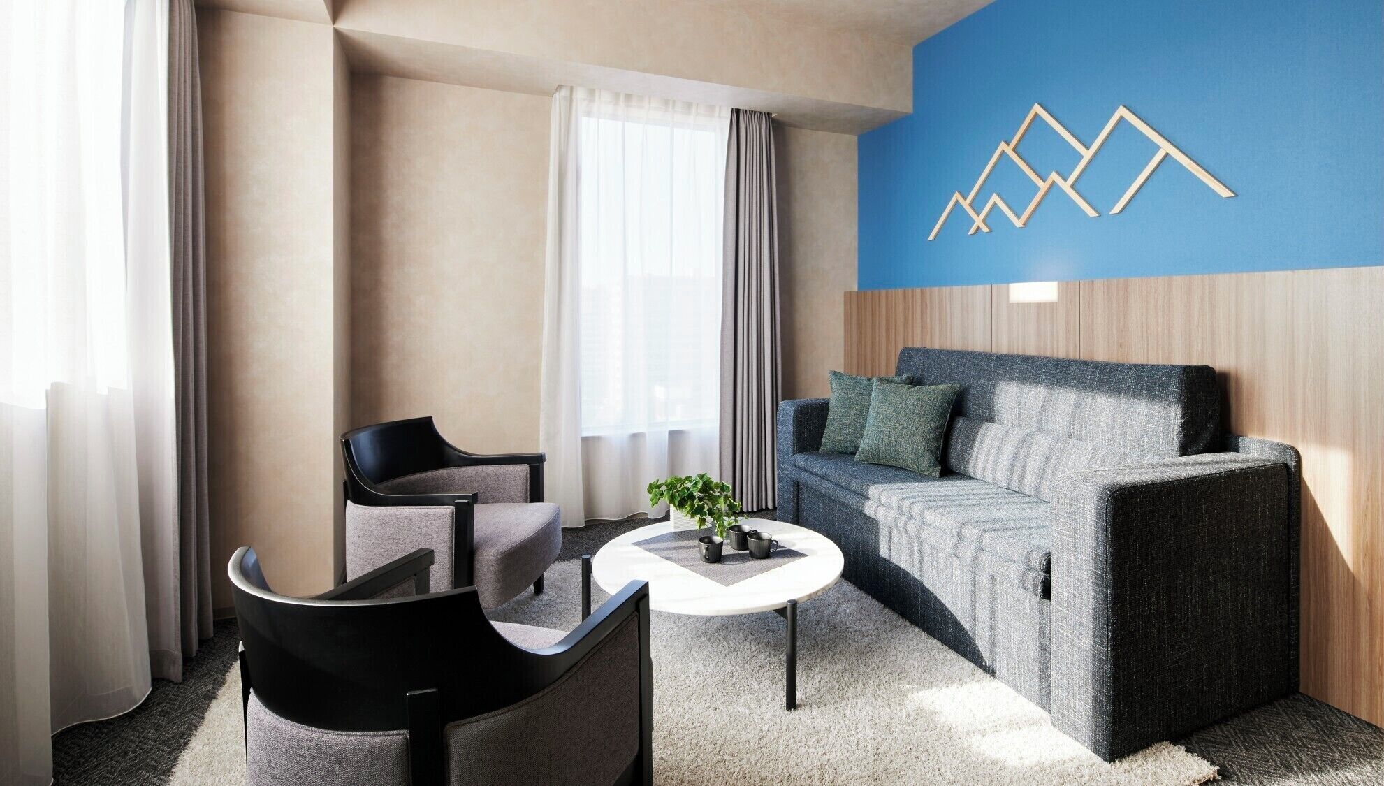 Una habitación de hotel moderna con un sofá gris, sillones negros, una mesa de centro blanca y una pared azul con diseño geométrico.