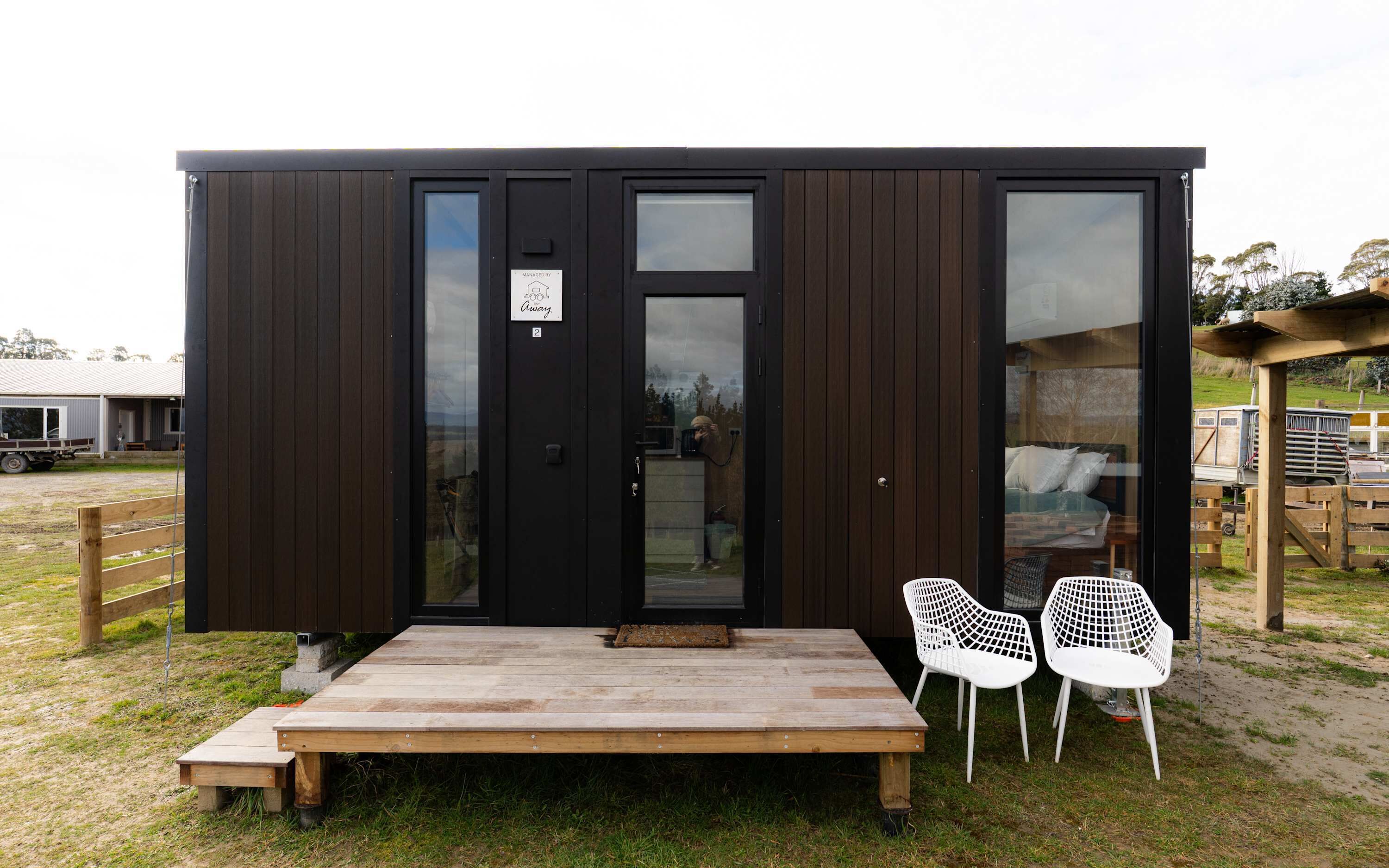 Een moderne, donker houten blokhut met een grote schuifdeur, een klein terras met een bankje en twee witte plastic stoelen.