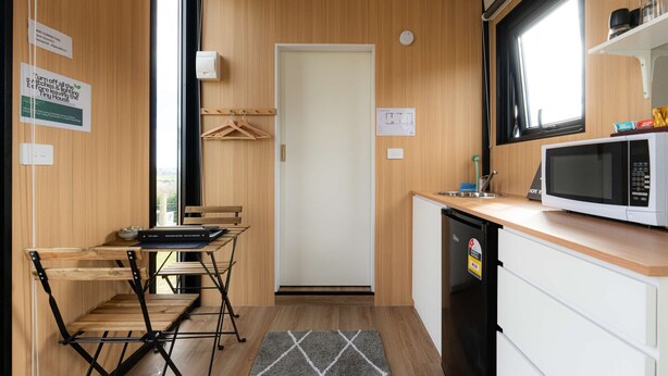 Een kleine keuken met een magnetron, spoelbak en houten keukenkastjes. Er is een zithoekje met een tafel en stoelen.