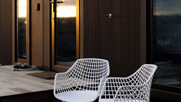 Een modern gebouw met een gevel van grote glazen deuren en ramen, een houten terras en twee witte metalen stoelen.