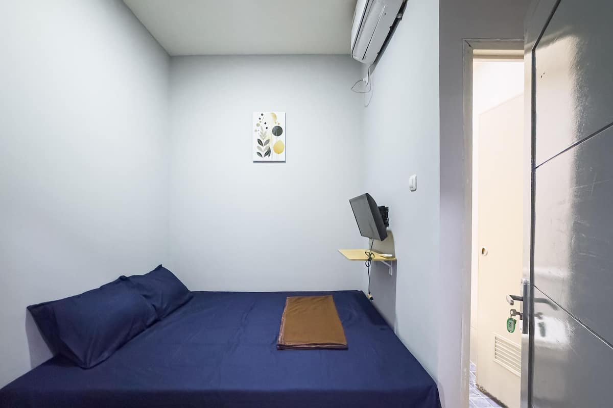 A small, single-bed room with a blue bedspread, a brown blanket, a wall-mounted TV, and a small picture.