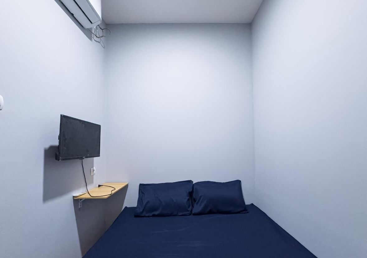 A small, single-bed room with a dark blue bedspread, a mounted flat-screen TV on a wooden shelf, and a white wall.