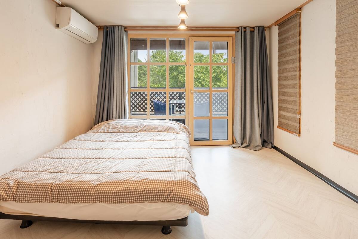 A bedroom with a bed, a window with curtains, a wall-mounted air conditioner, and a view of greenery outside.