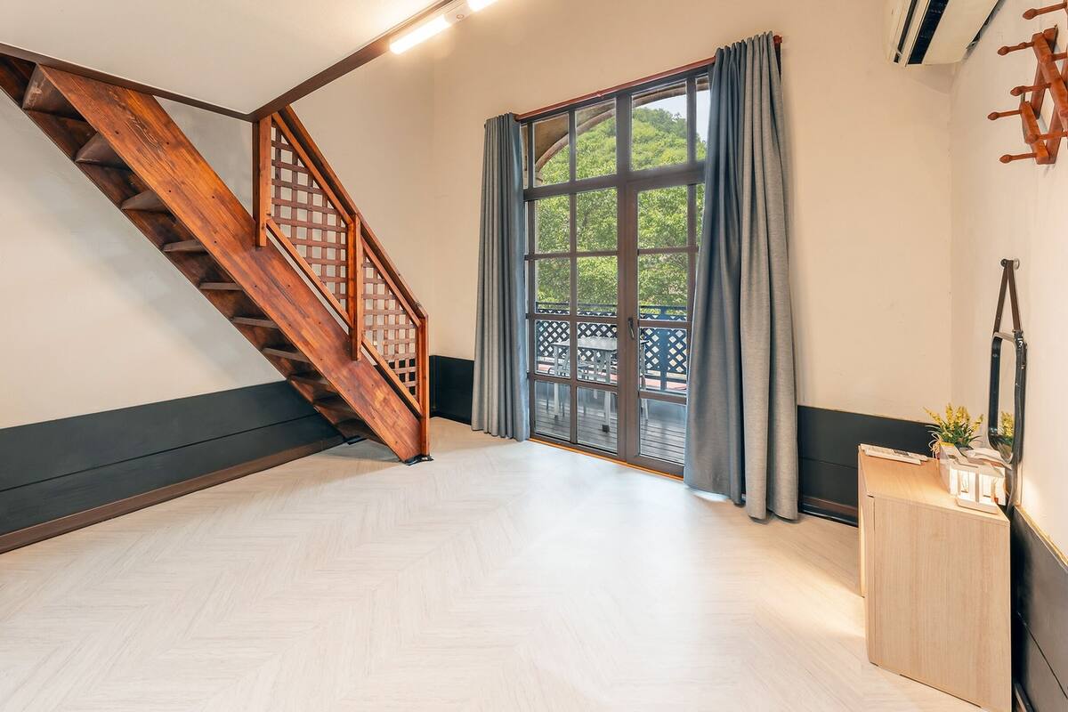 A spacious room with a wooden staircase, a large window with curtains, and a herringbone-patterned floor.