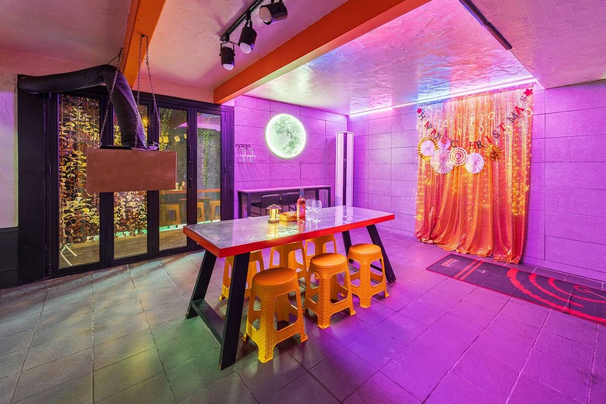 A modern interior with a glass table and orange stools, a purple-lit wall, and a ceiling with a light fixture.