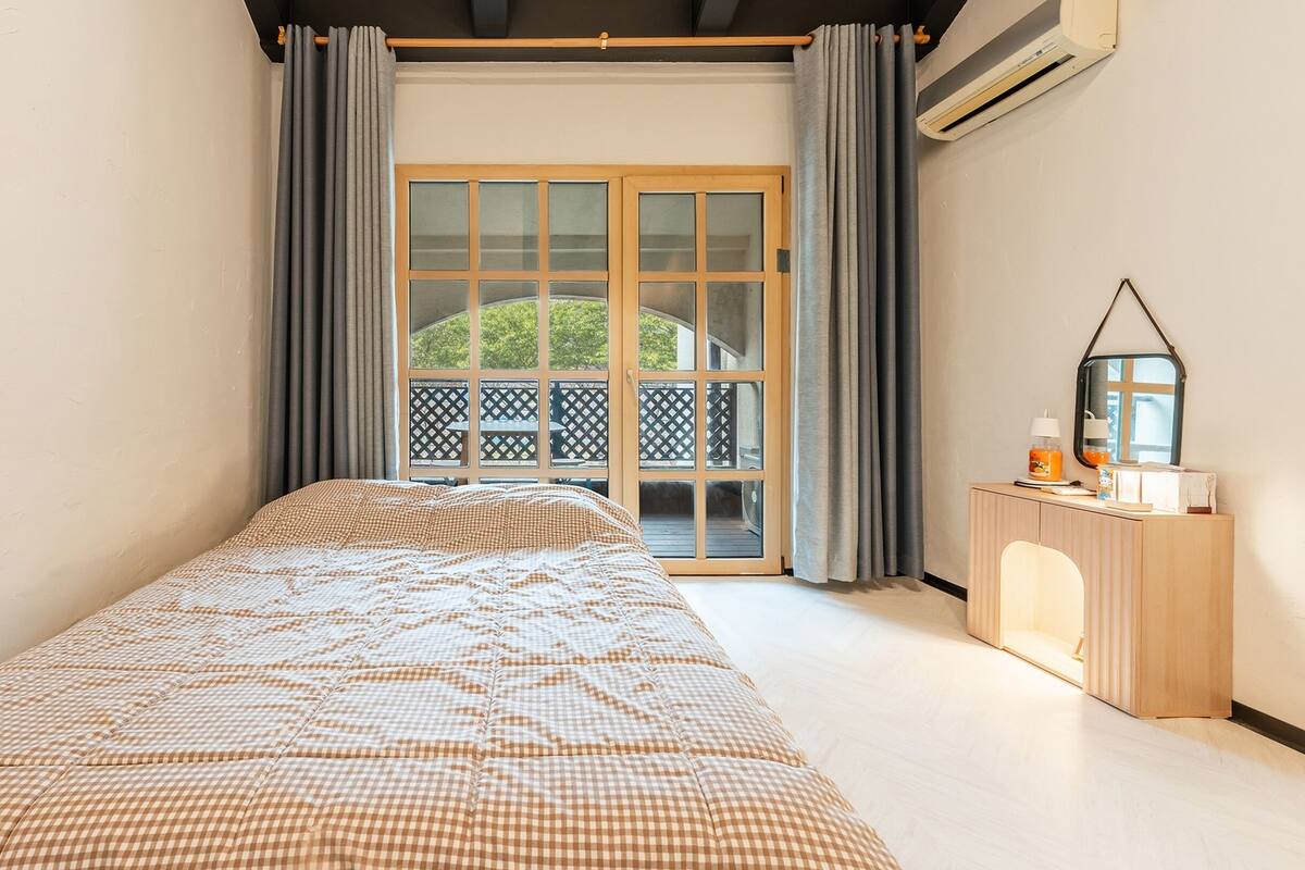 A bedroom with a checkered bedspread, a wooden bedside table with a lamp, and a view of a balcony with a lattice door.