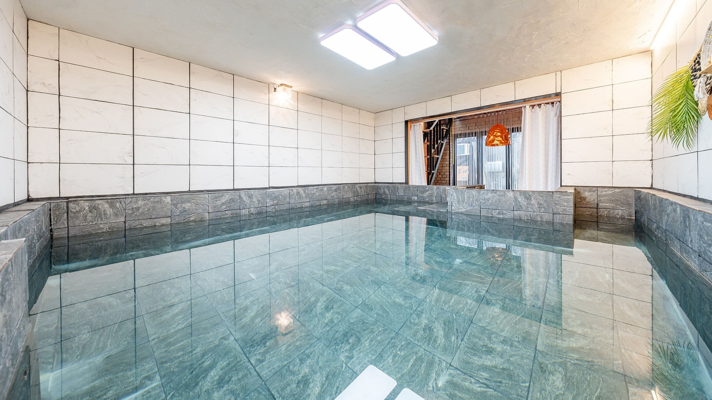 Una piscina interior amplia con superficie de agua cristalina, rodeada de paredes de azulejos y un borde de mármol.