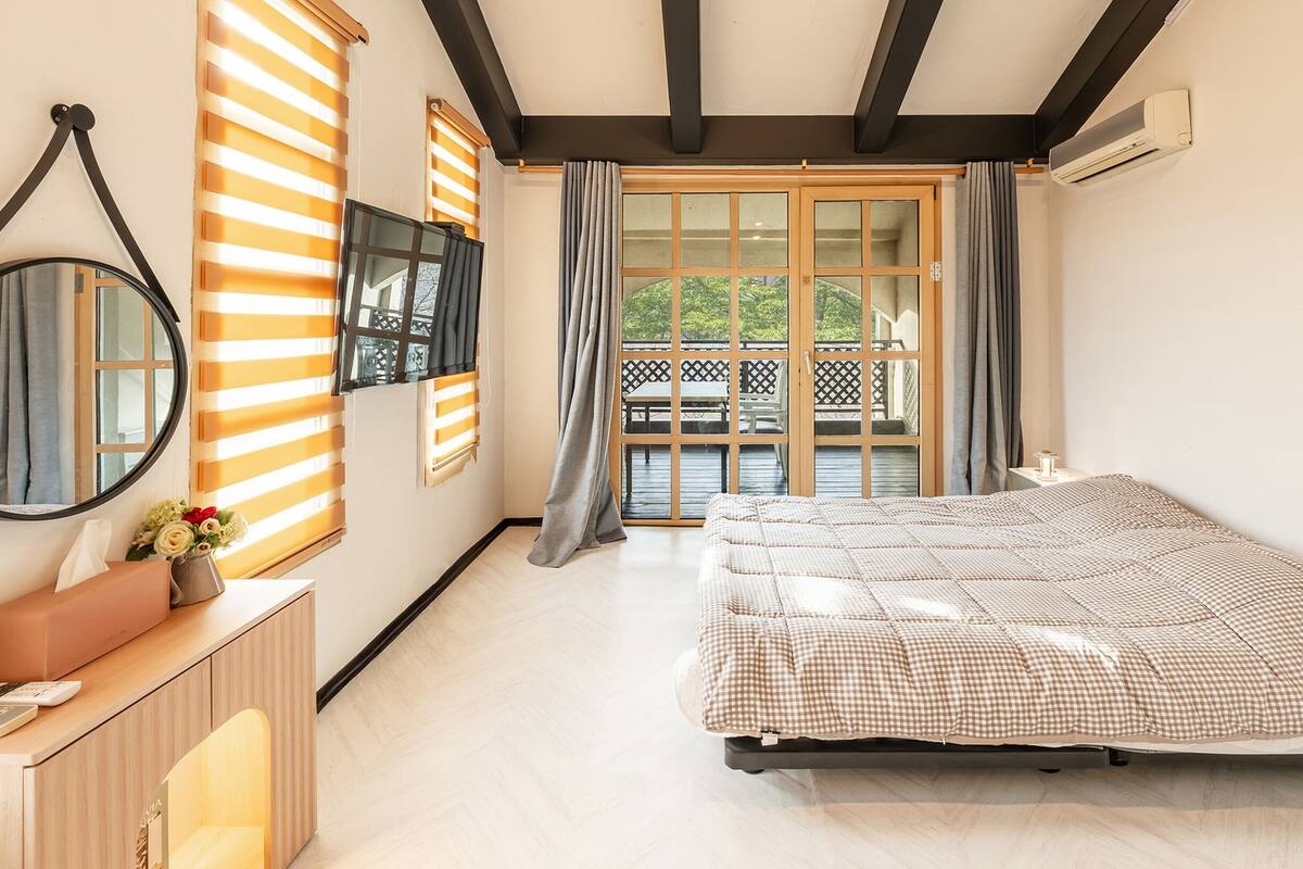 A bedroom with a bed, a desk, a mirror, and a view of the outdoors.