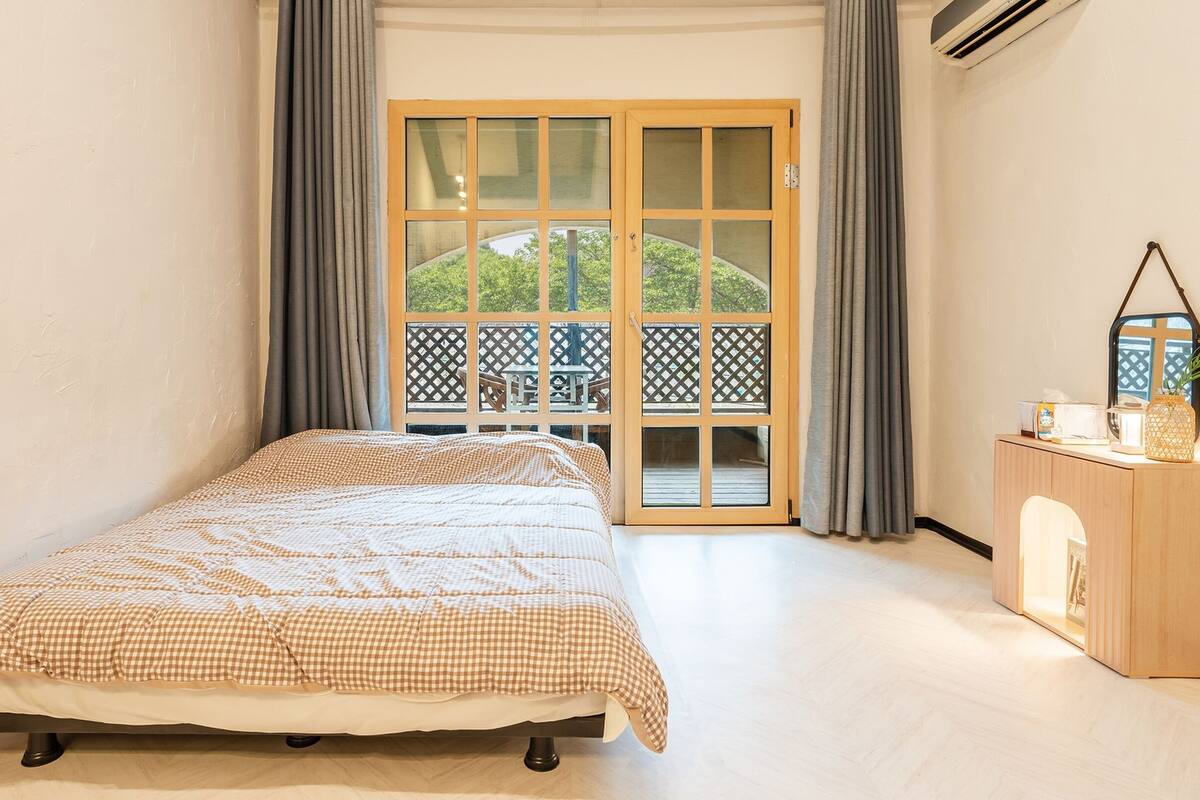 A bedroom with a bed, a wooden dresser, a mirror, and a view of a balcony with a table and chairs.