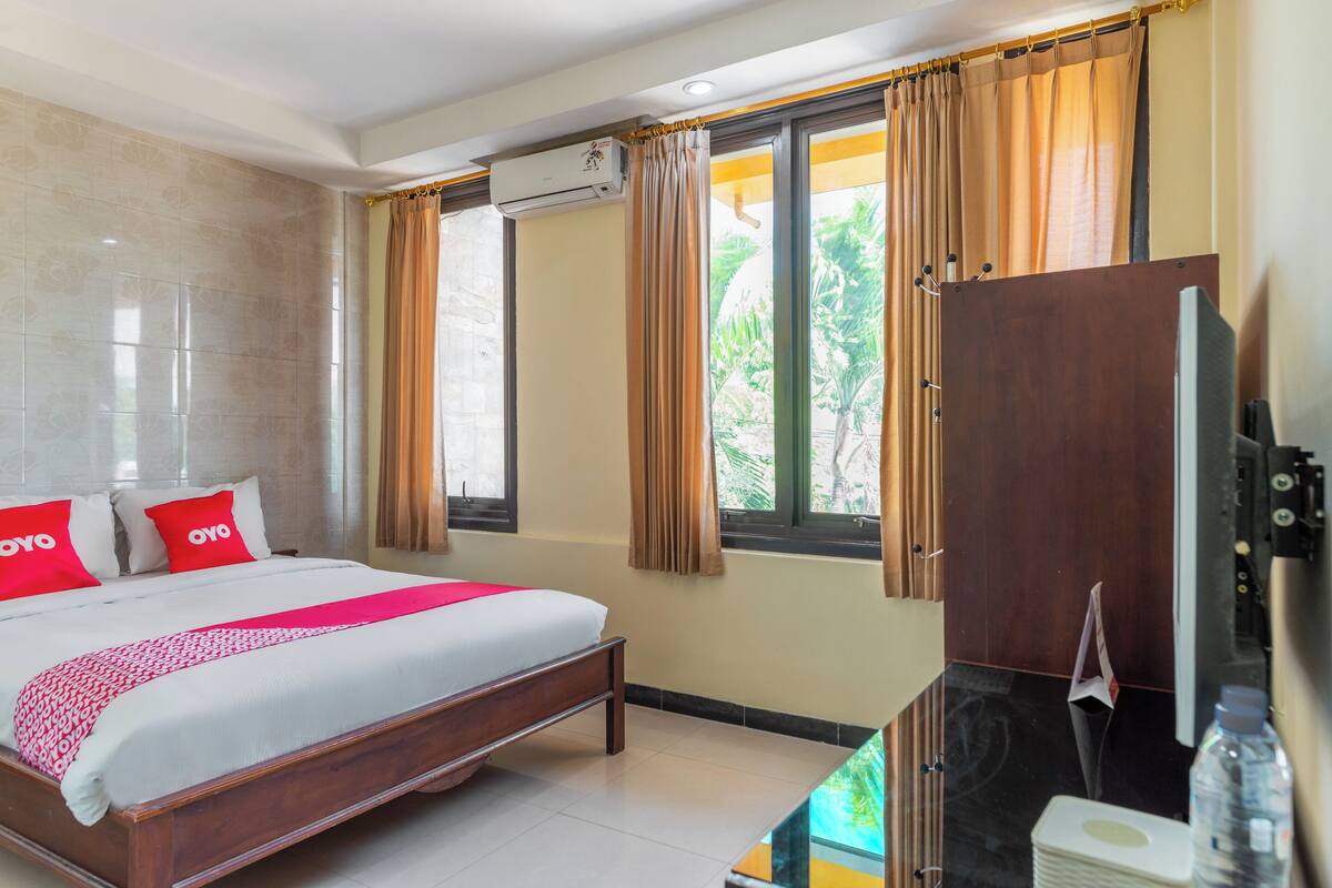 A hotel room with a bed, a television, a window with curtains, and a wall-mounted air conditioning unit.