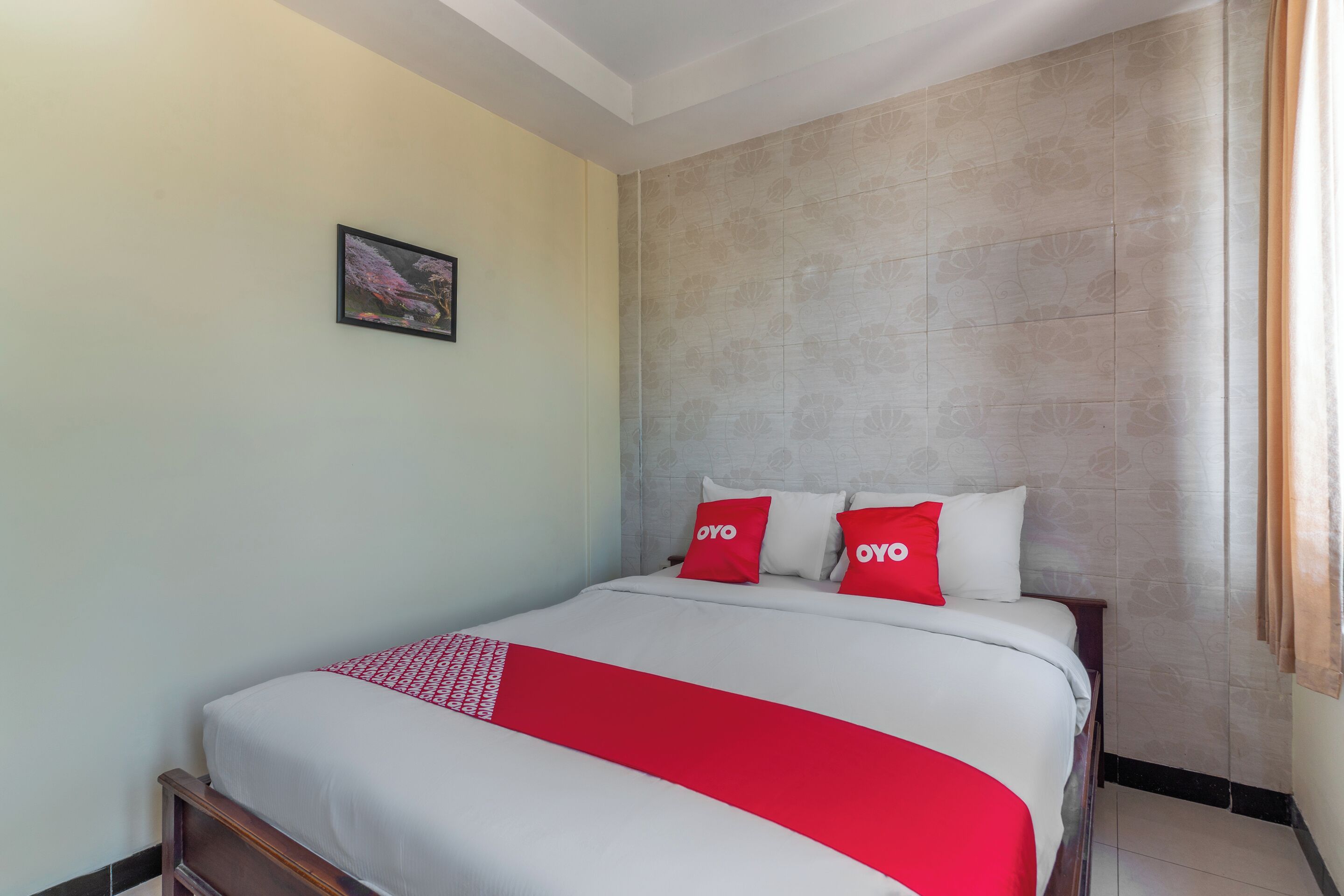 Een net opgemaakt bed met een rode en witte spreidek, twee kussens met 'OYO'-branding, een ingelijst beeld aan de muur, en een patroon op de achterwand.