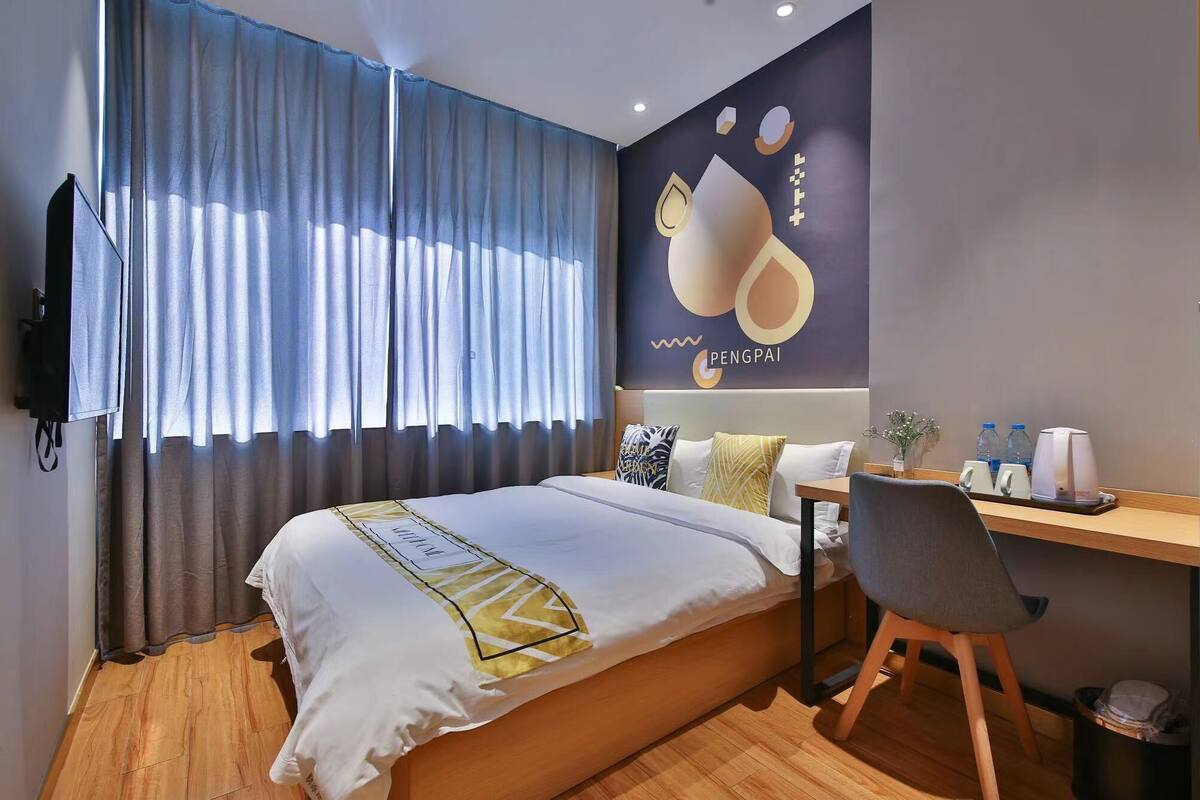 A hotel room with a bed, a desk, a chair, a TV, and a wall with a logo.