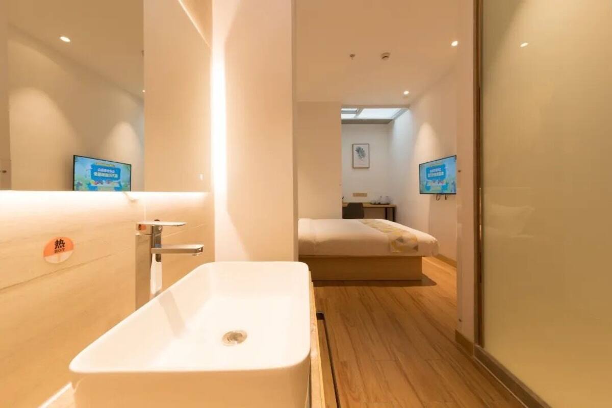 A modern bathroom with a large white sink, a mirror, and a wooden floor. There is a bed visible in the background.