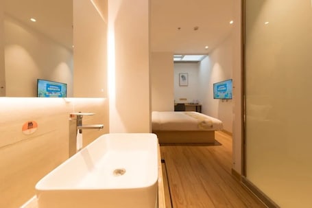 A modern bathroom with a large white sink, a mirror, and a wooden floor. There is a bed visible in the background.