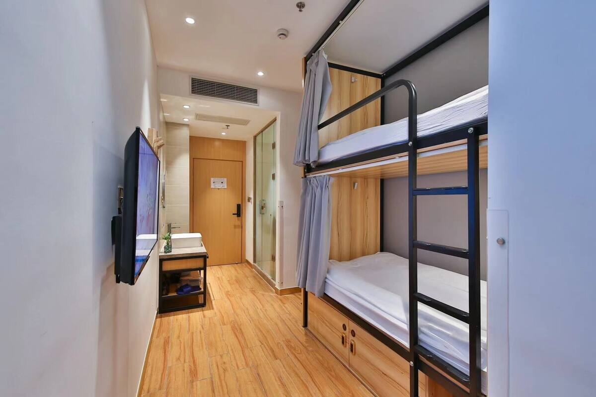 A narrow hotel room with a bunk bed, a small desk with a mirror, and a television.