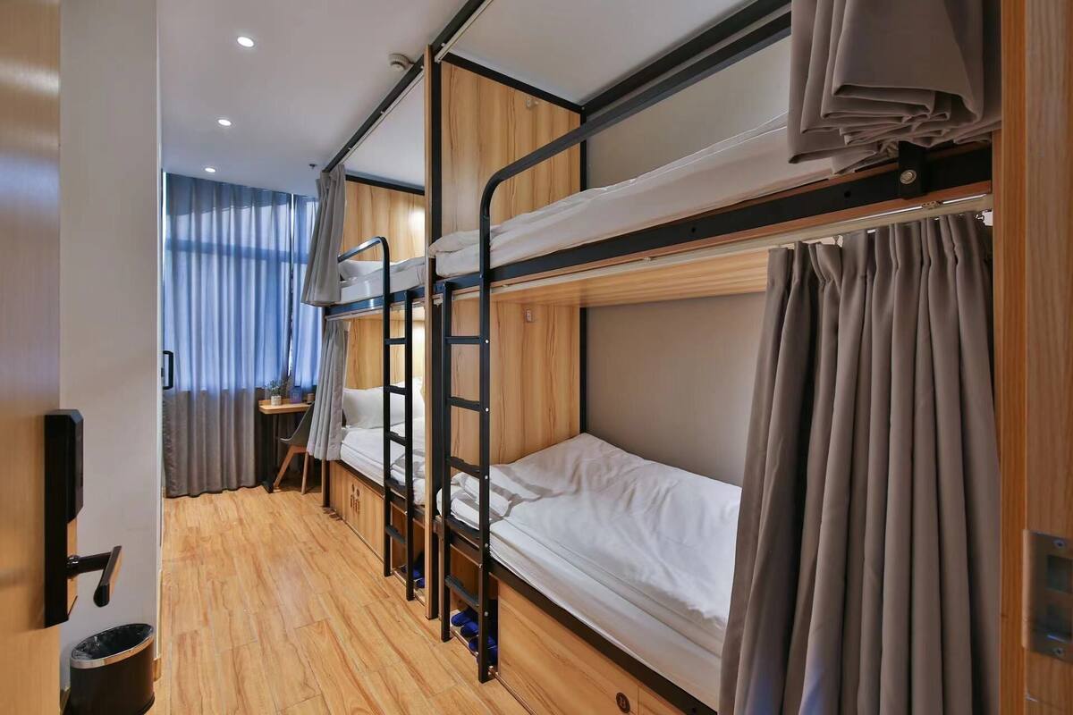 A bunk bed room with wooden flooring, a desk with a chair, and a window with curtains.