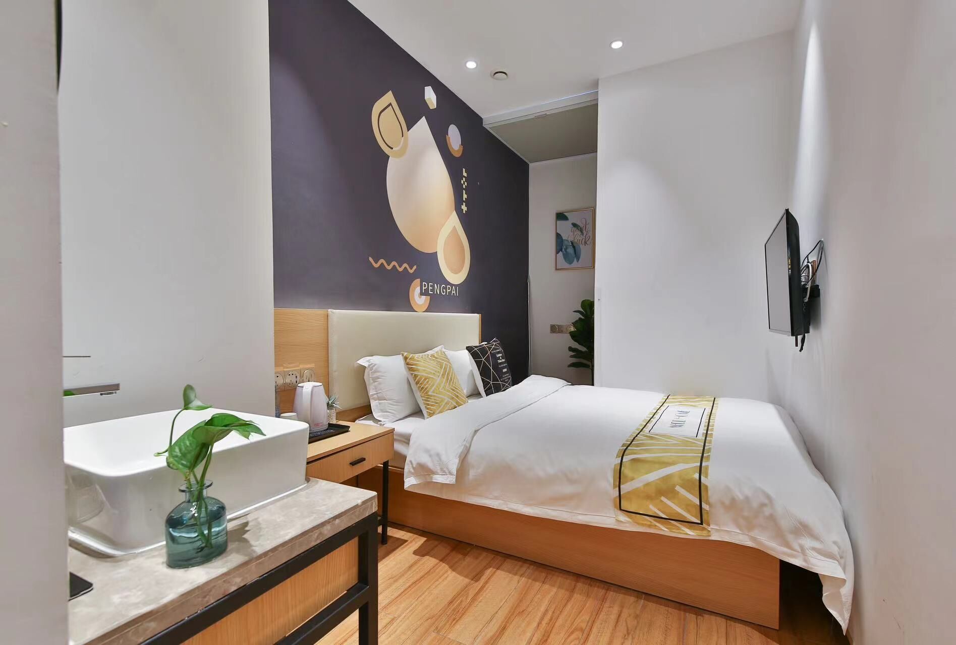 A modern hotel room with a bed, a bedside table with a lamp, a wall-mounted TV, and a large abstract artwork.