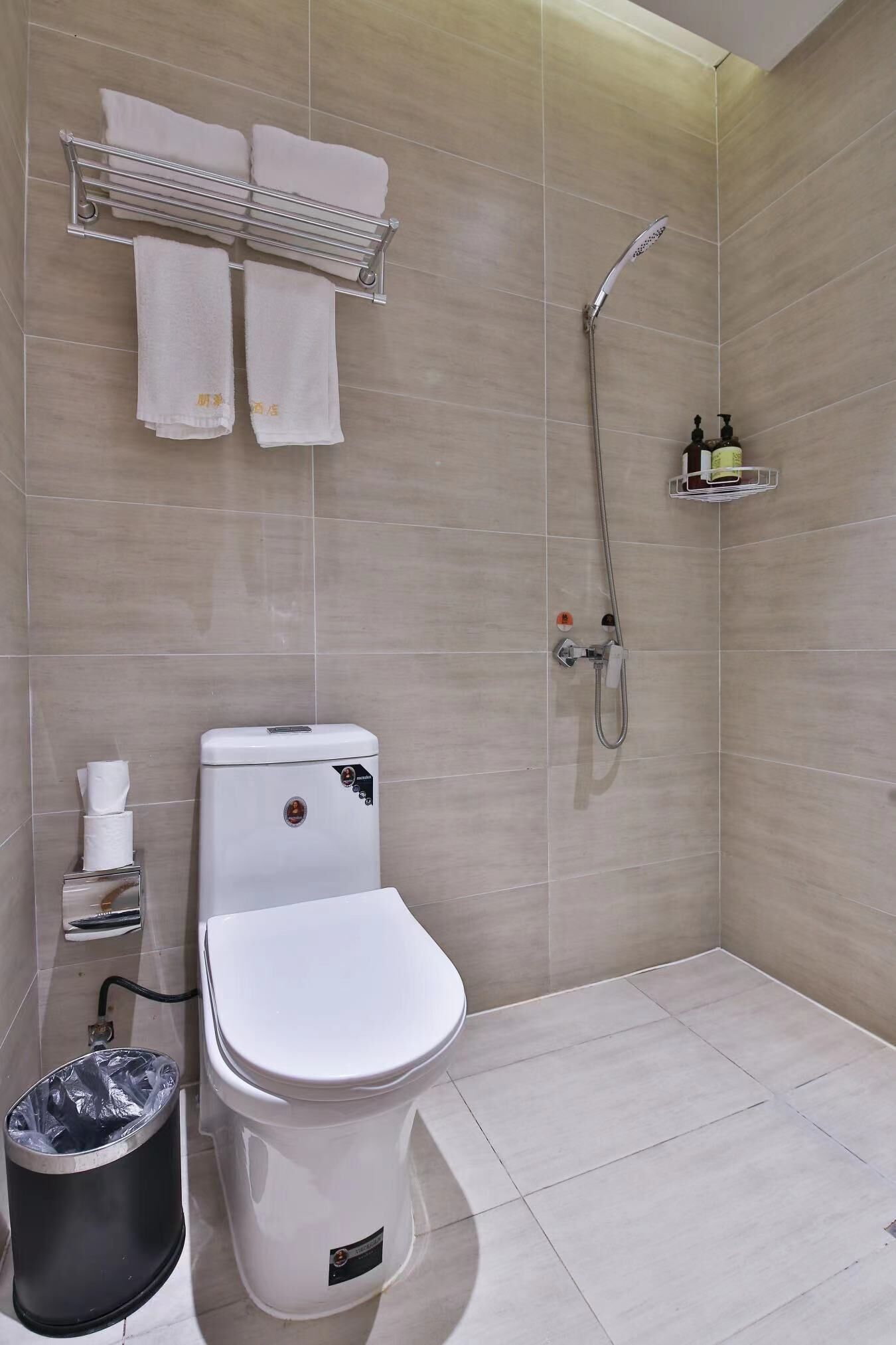 Ein modernes Badezimmer mit Toilette, einem Mülleimer, einem Handtuchhalter mit Handtüchern, einer Dusche und einem kleinen Ablagebereich für Pflegeprodukte.