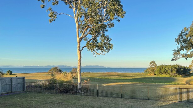 A grassy area with a fence, a tree, and a view of the ocean and mountains.