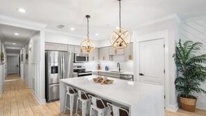 A modern kitchen with stainless steel appliances, a large island with bar stools, pendant lighting, and a potted plant.