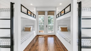 A room with two bunk beds, wooden flooring, white walls, and a balcony with a wooden railing.