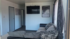 A hotel room with a sofa, a flat-screen TV, framed artwork, and a door leading to another room. - Cozy Cove Ohope Beach (Ohope)